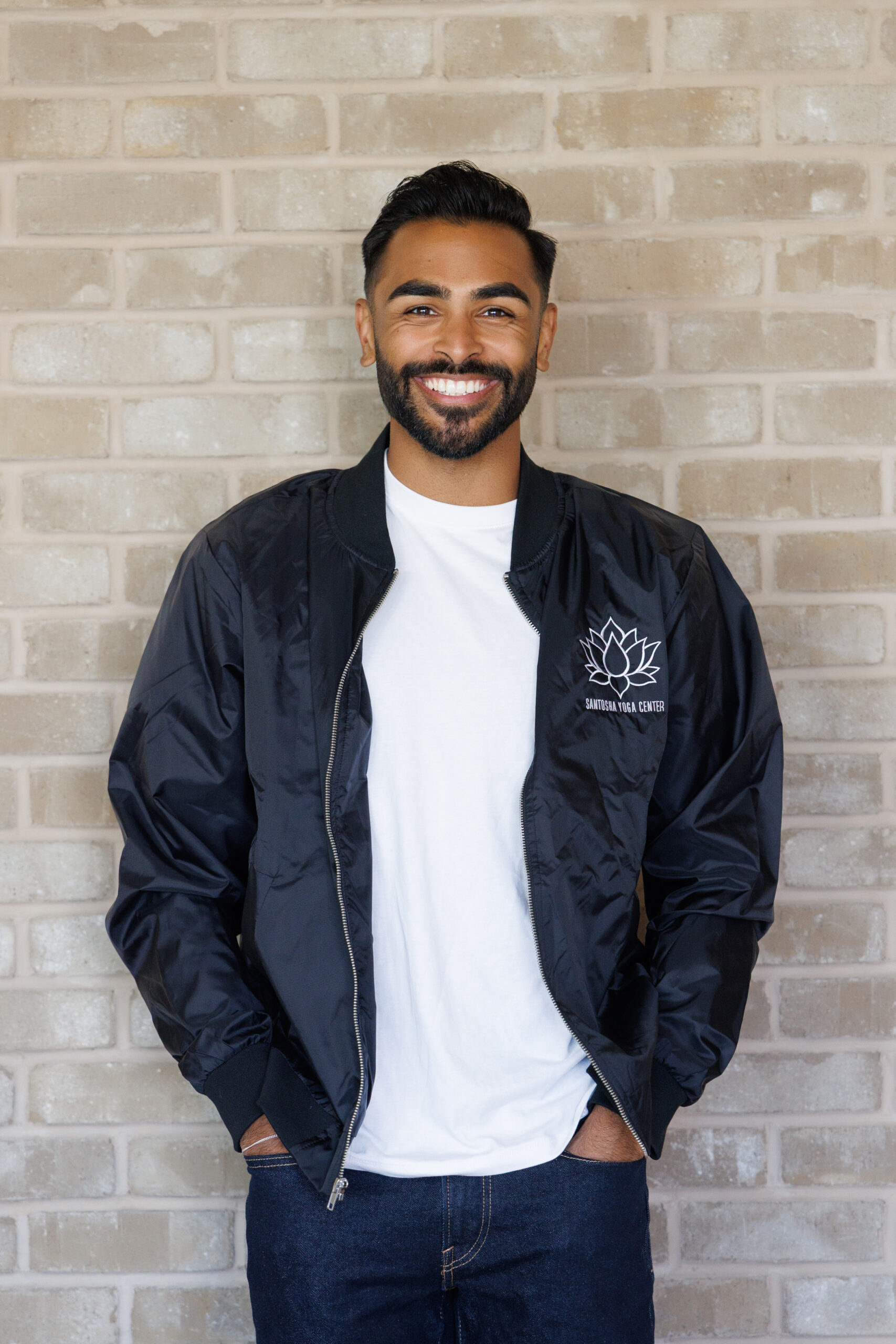 Smiling man in a black bomber jacket featuring the "Santosha Yoga Center" logo, posed against a brick wall. Reflects a welcoming atmosphere.