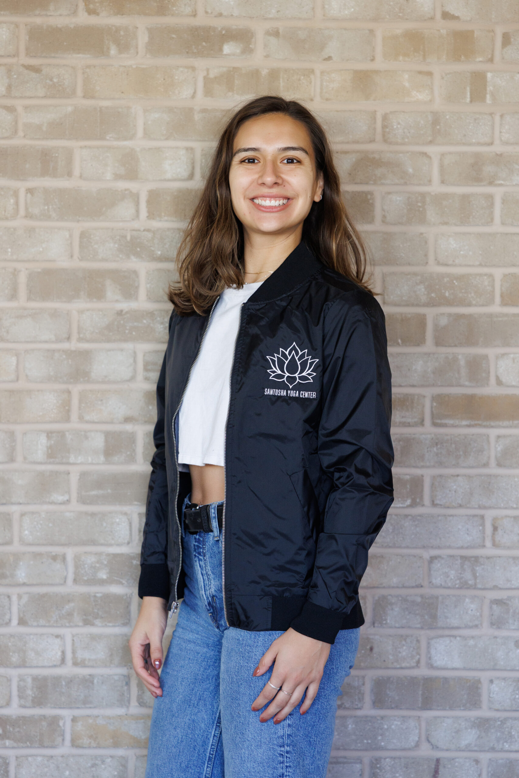 Smiling woman in a black bomber jacket with a lotus logo, standing against a textured brick wall, representing Santushia Yoga Center.