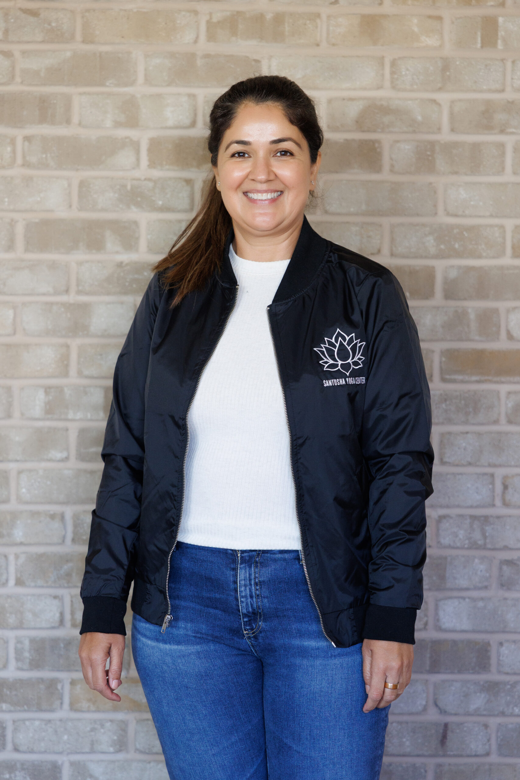 Smiling woman in a black bomber jacket with a lotus logo, posed against a textured brick wall, representing a friendly and approachable vibe.