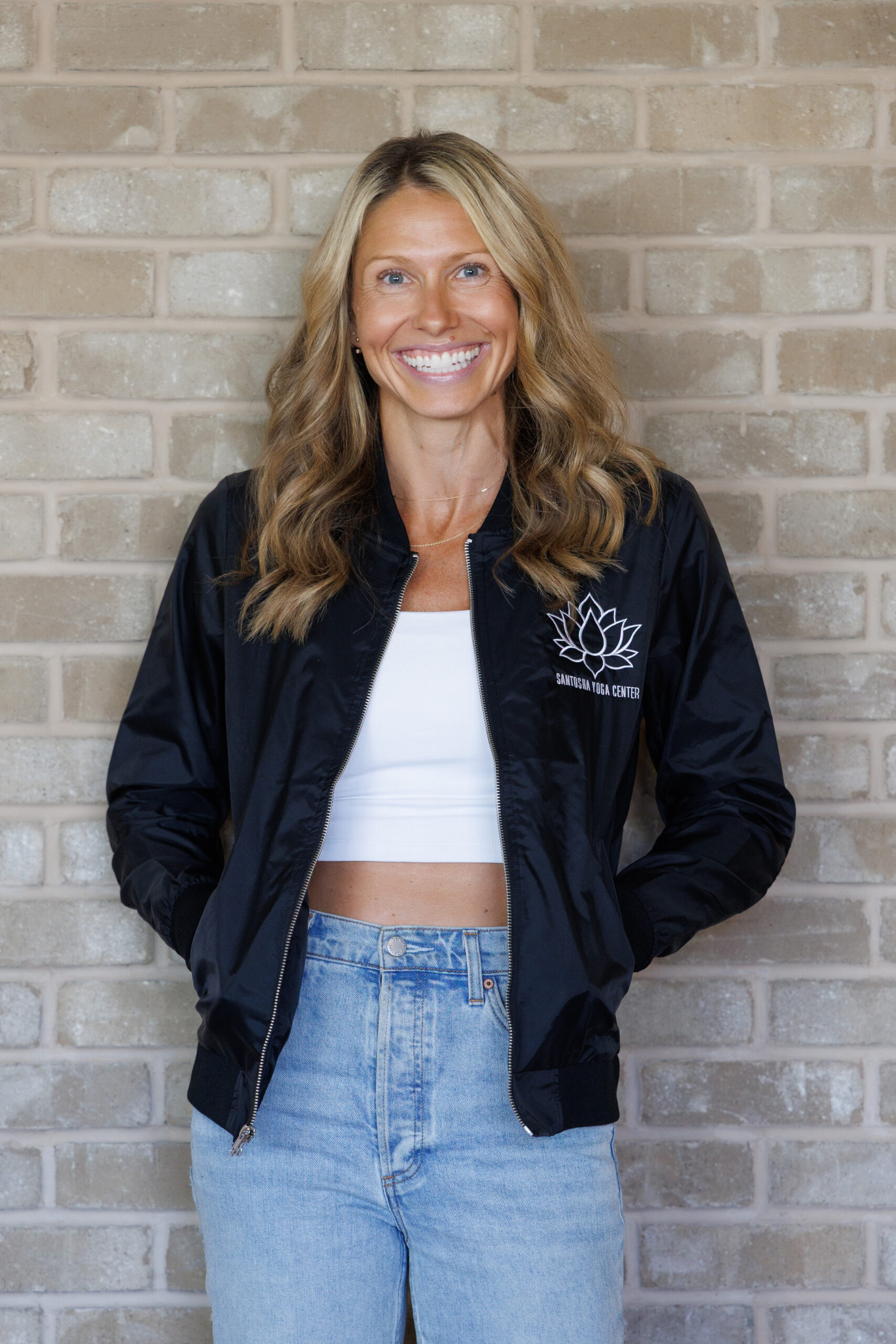 Smiling woman in a black bomber jacket with a lotus logo, standing against a brick wall, promoting a yoga center.