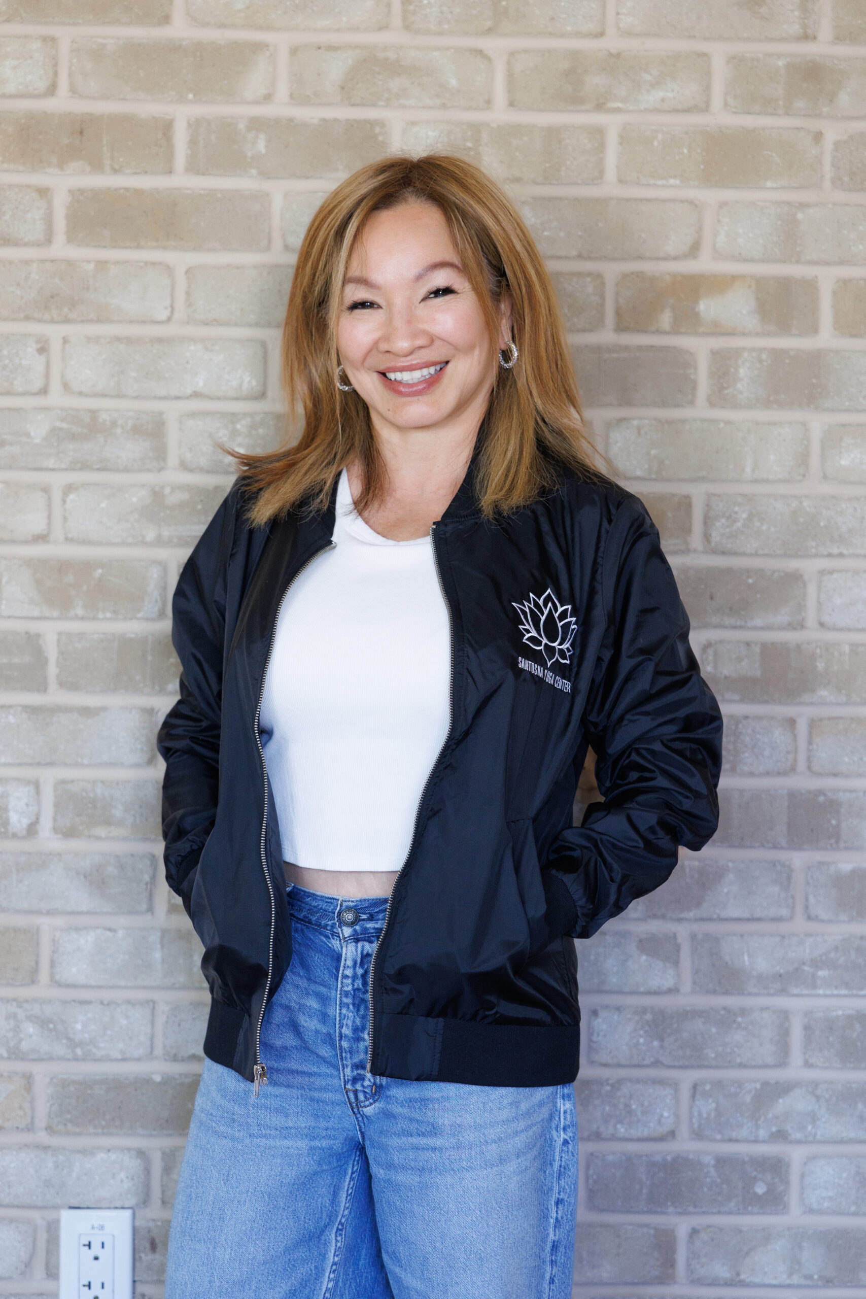 A woman with long, light brown hair smiles confidently in a black bomber jacket and white top, standing in front of a textured brick wall.