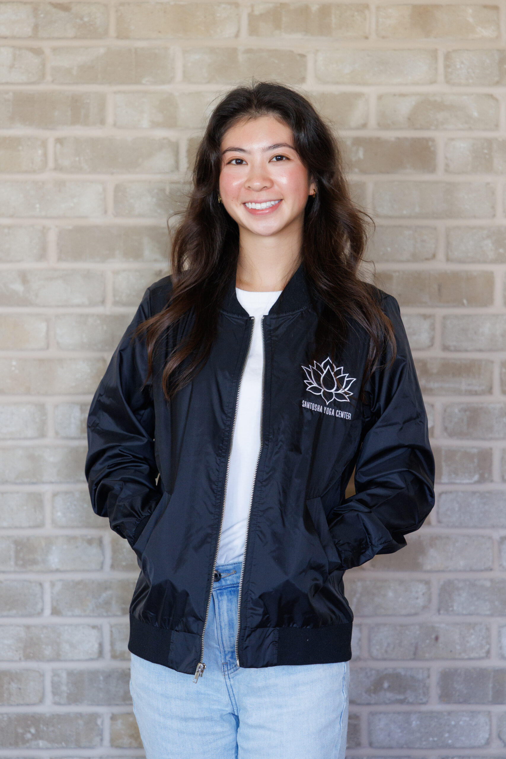 A young woman smiles in a black bomber jacket featuring the Santosha Yoga Center logo, standing against a textured brick wall.