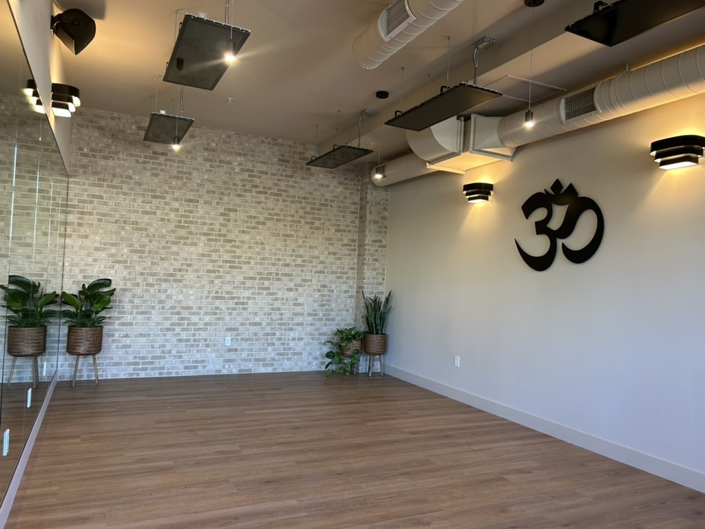 Yoga studio interior featuring a wooden floor, brick wall, and potted plants. The symbol "Om" is displayed prominently, signifying a peaceful atmosphere.